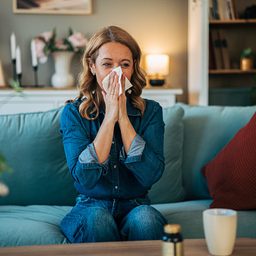 Woman with blonde hair sitting on a sofa in a living room, blowing her nose with a tissue while feeling unwell from allergies.