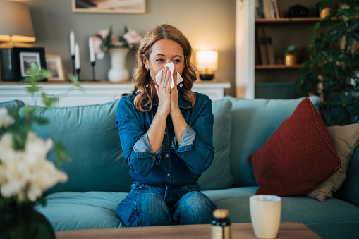 Woman with blonde hair sitting on a sofa in a living room, blowing her nose with a tissue while feeling unwell from allergies.